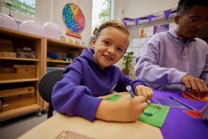 Een foto van paarse vrijdag in een klaslokaal op een basisschool. Er hangen paarse vlaggetjes, kinderen dragen paarse kleren. Een jongetje maakt een tekening aan tafel en kijkt blij de camera in.