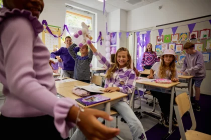 Een foto van paarse vrijdag in een klaslokaal op een basisschool. Er hangen paarse vlaggetjes, kinderen dragen paarse kleren. Op de voorgrond staat een lerares met haar rug naar de camera. Een kind poseert blij met de handen omhoog.