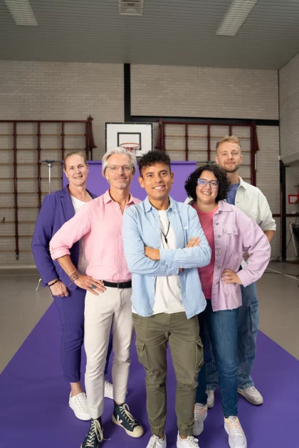 Een groepje leraren staat in de gymzaal van een school. Ze poseren voor een groot paars scherm, en kijken lachend de camera in.