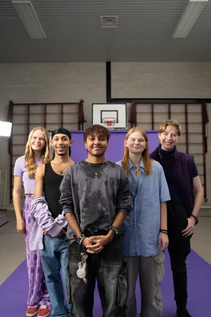 Een groepje leerlingen staat in de gymzaal van een school. Ze poseren voor een groot paars scherm, en kijken lachend de camera in.