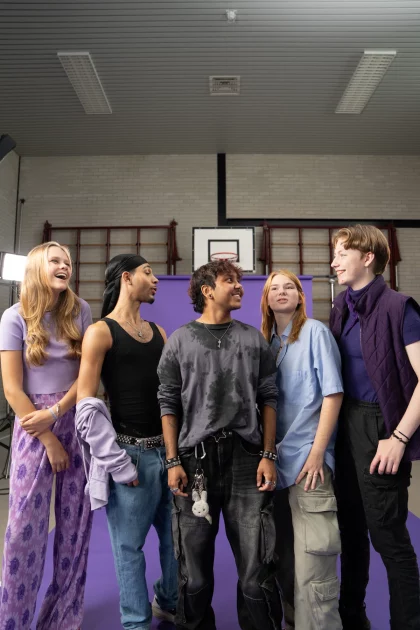Een groepje leerlingen staat in de gymzaal van een school. Ze poseren voor een groot paars scherm, en kijken lachend de camera in.