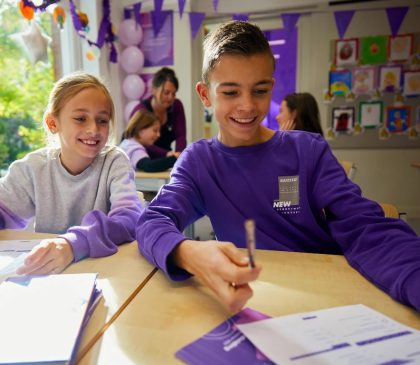 Een foto van paarse vrijdag in een klaslokaal op een basisschool. Er hangen paarse vlaggetjes, kinderen dragen paarse kleren. Een jongen en een meisje zijn op de voorgrond naast elkaar aan het werk in de schoolbanken, ze schrijven iets op papier en lachen allebei.