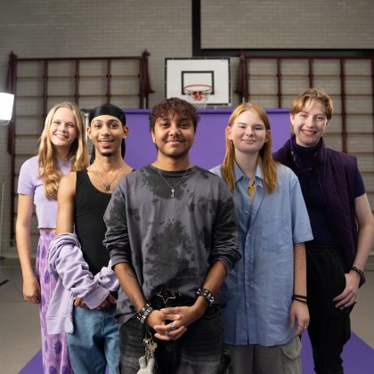 Een groepje leerlingen staat in de gymzaal van een school. Ze poseren voor een groot paars scherm, en kijken lachend de camera in.