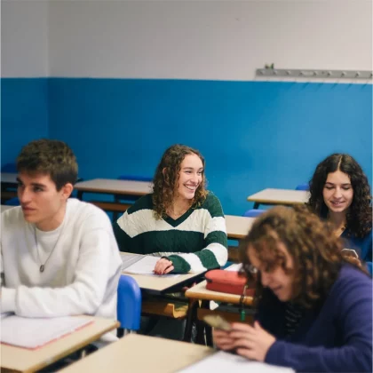 Foto van vier middelbare scholieren in de schoolbanken met opengeslagen lesboeken. Twee meisjes lachen naar elkaar. Een jongen voorin kijkt vooruit, een meisje voorin kijkt op haar mobiel.