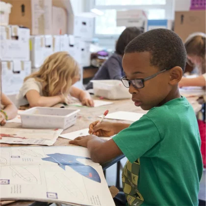 Een foto van een jongetje uit de onderbouw van de basisschool in een groen shirt aan een tafel in een klaslokaal. Hij kijkt in een boek en schrijft iets op een papier. Achter hem zitten meerdere kinderen te werken.