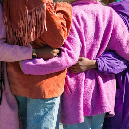 Een foto van ruggen van een aantal mensen die de armen om elkaar hebben geslagen. Ze dragen paarse, roze en oranje kleren.